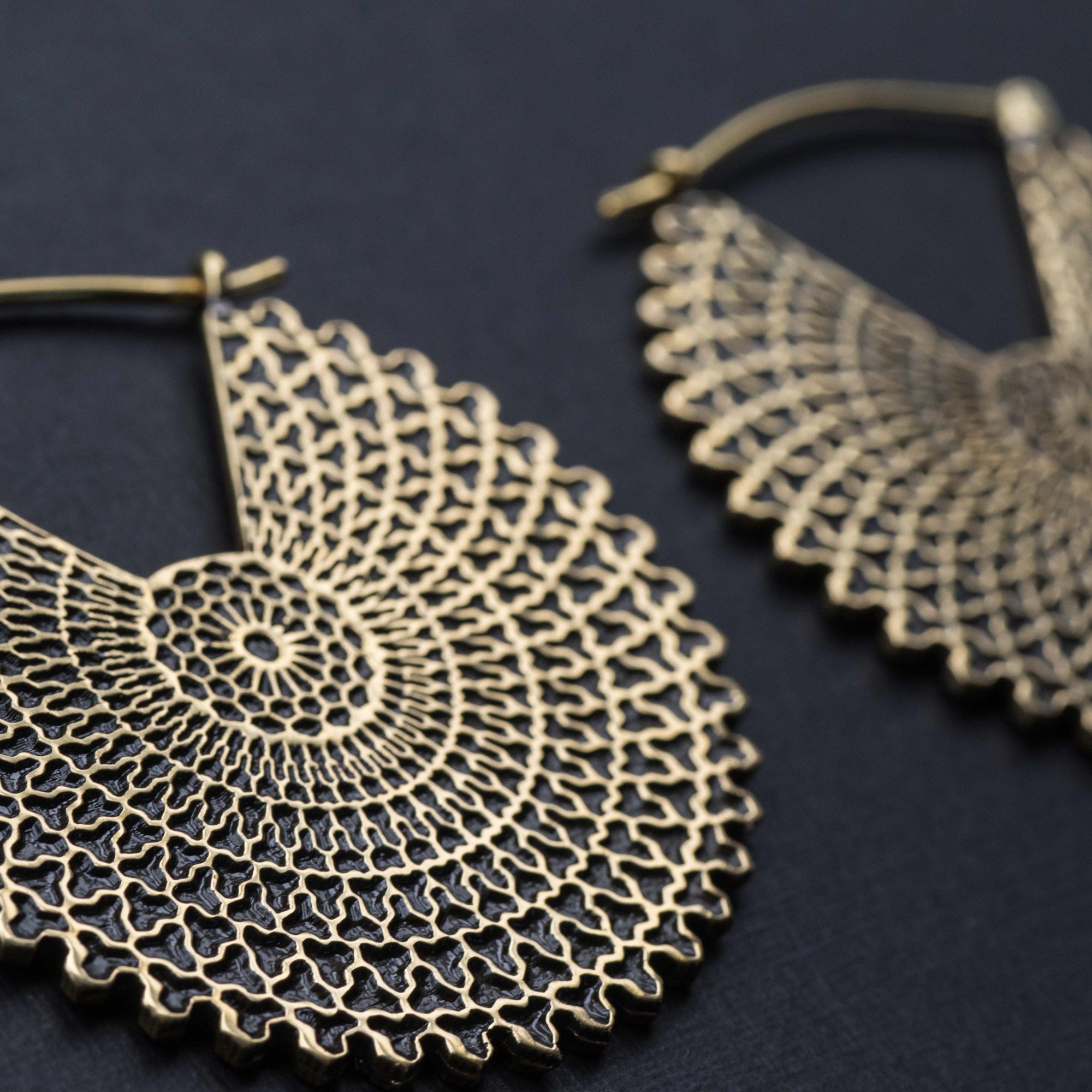 Close-up of a pair of VORTEX Tribal Oversized Mandala Disc Hoop Earrings in Gold by Kaya Movement. These 16 gauge earrings feature a lace-like geometric pattern with concentric loops and tiny cross shapes, set against a dark background, emphasizing their delicate design and metallic sheen.